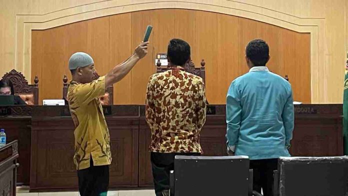 Prosesi pengambilan sumpah saksi di ruang sidang; seorang pria berbaju batik berdiri tegak sementara petugas pengadilan mengangkat Al-Qur'an di atas kepalanya.