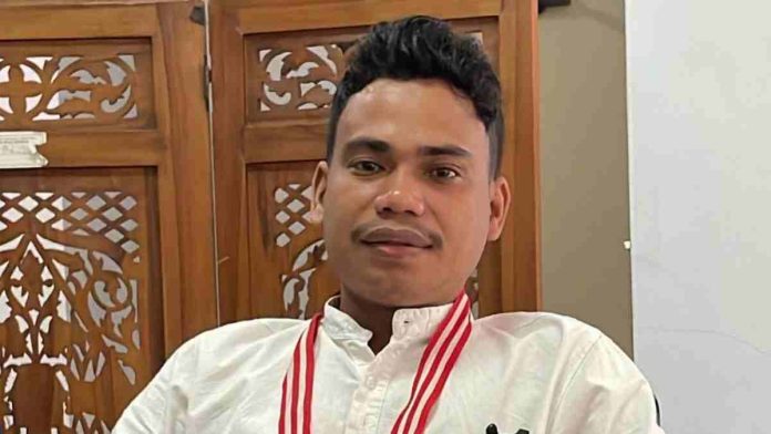 Foto close-up seorang pemuda berambut hitam pendek bergelombang, mengenakan kemeja putih dengan aksen tali kalung merah-putih di leher. Ia berpose menghadap kamera dengan latar belakang pembatas ruangan kayu berukir khas tradisional.