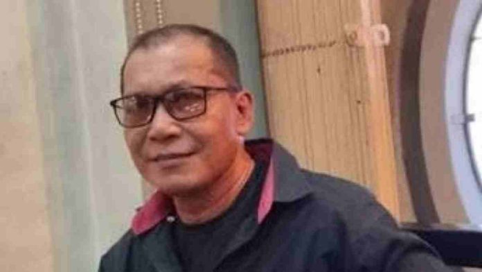 Foto close-up seorang pria paruh baya berkacamata, berambut pendek, dan berkumis tipis. Pria tersebut mengenakan kemeja ganda berwarna hitam dengan kerah bagian dalam berwarna merah muda/merah tua. Ia menatap kamera dengan ekspresi tersenyum tipis. Latar belakang buram menunjukkan elemen interior ruangan berwarna cokelat.