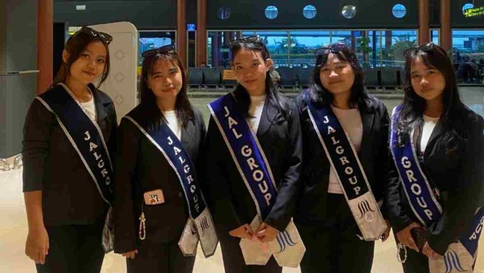 Lima wanita muda berdiri berjajar mengenakan seragam jas hitam dan kaus putih. Masing-masing mengenakan selempang biru tua bertuliskan 