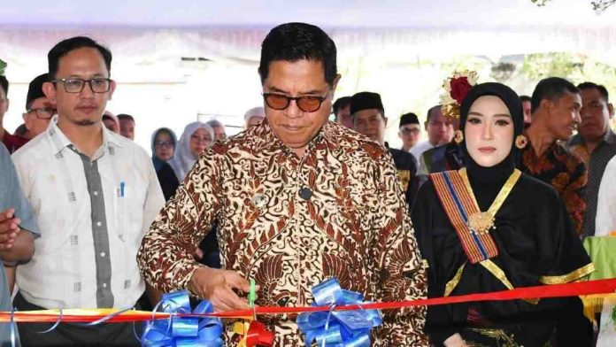 Bupati Lombok Timur mengenakan batik cokelat dan kacamata sedang memotong pita merah dalam acara peresmian. Di sampingnya berdiri seorang wanita mengenakan pakaian adat Sasak hitam dengan selempang dan hiasan bunga, serta seorang pria berkacamata mengenakan kemeja putih.