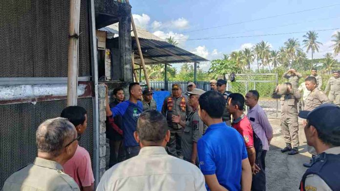 Sejumlah petugas Satpol PP dan pejabat daerah sedang berdialog dengan pengelola peternakan di depan sebuah bangunan kandang ayam. Para petugas mengenakan seragam dinas, sementara pengelola menggunakan kaus biru. Suasana luar ruangan di bawah cuaca cerah dengan latar pohon kelapa.
