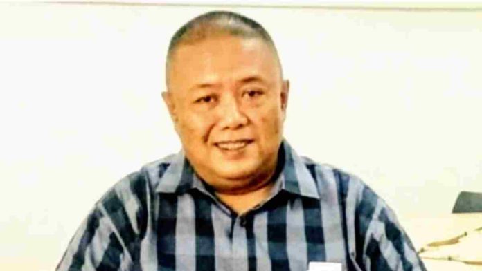 Foto close-up Direktur Mi6, Bambang Mei Finarwanto, atau akrab disapa Didu. Ia adalah seorang pria paruh baya berkepala plontos dan tersenyum ramah ke arah kamera. Didu mengenakan kemeja lengan pendek bermotif kotak-kotak besar berwarna kombinasi biru tua, hitam, dan putih. Latar belakang foto berwarna putih polos dan agak buram.