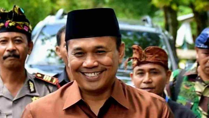 Foto close-up Dr. H. Ahsanul Khalik yang sedang tersenyum lebar mengenakan kemeja cokelat dan peci hitam. Di latar belakang tampak beberapa pria lain, termasuk seorang petugas kepolisian berseragam dan warga yang mengenakan udeng (ikat kepala khas Bali/Lombok).
