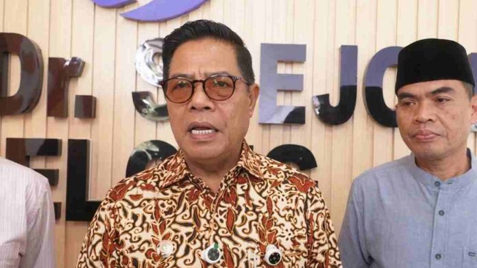 Foto jarak dekat (close-up) Bupati Lombok Timur, Haji Khairul Warisin, mengenakan kemeja batik bermotif cokelat dan kacamata berbingkai gelap. Ia tampak sedang berbicara memberikan keterangan pers di depan latar belakang dinding kayu bertuliskan 