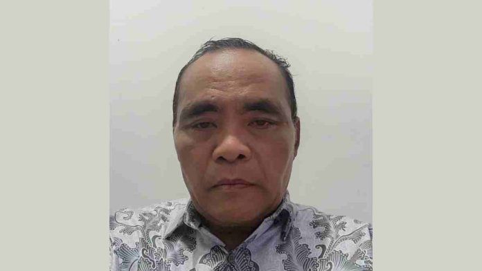 Foto close-up Dr. H. Lalu Moh. Fahri, MH mengenakan kemeja batik bermotif flora dengan latar belakang dinding putih polos, menatap ke arah kamera dengan ekspresi formal.