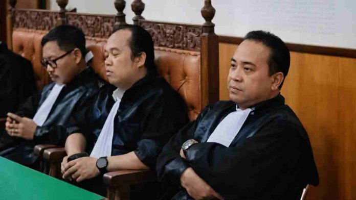Tiga orang pria mengenakan toga advokat berwarna hitam dengan kerah putih (jabot) duduk di kursi kayu berukir di dalam ruang sidang. Pria paling depan tampak melipat tangan di dada dengan ekspresi serius menyimak persidangan, sementara dua rekan di sampingnya juga duduk dalam posisi mendengarkan di balik meja hijau.