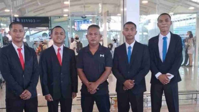 Lima orang pria berfoto bersama di dalam terminal Bandara Internasional Lombok. Pria di tengah mengenakan kaos polo hitam bertanda logo H. Di sisi kirinya terdapat dua pria muda mengenakan jas hitam dan dasi merah, sementara di sisi kanannya terdapat dua pria muda lain mengenakan jas hitam dan dasi biru. Mereka berpose formal menghadap kamera. Latar belakang menampilkan suasana bandara dengan dinding kaca dan layar informasi penerbangan yang kabur.