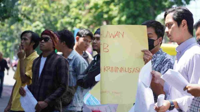 Sekelompok pemuda melakukan aksi demonstrasi di pinggir jalan yang teduh oleh pepohonan. Salah satu peserta aksi di barisan depan memegang poster besar berwarna kuning dengan tulisan tangan merah dan hitam yang mencantumkan kata 