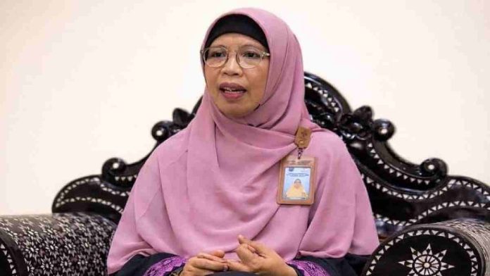 Seorang wanita berhijab pink dan berkacamata duduk di kursi ukir hitam. Ia mengenakan kartu identitas pegawai dan tampak sedang memberikan penjelasan dalam sesi wawancara.
