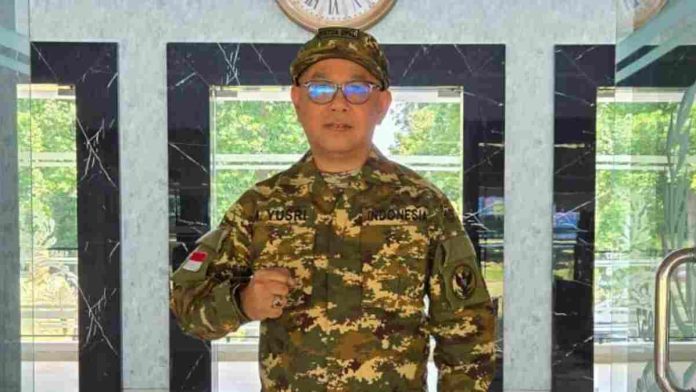 Muhammad Yusri mengenakan seragam loreng militer lengkap berdiri tegak di lobi gedung yang megah. Ia berpose dengan satu tangan mengepal di depan dada.