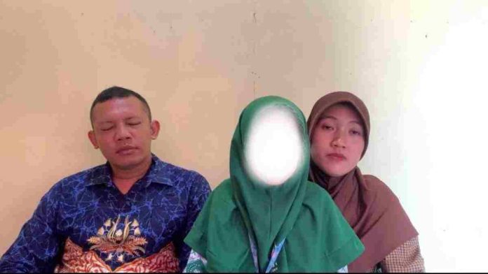 Pasangan suami istri duduk berdampingan dengan ekspresi sedih. Sang ayah mengenakan batik biru, sementara ibu berhijab cokelat merangkul anak mereka yang wajahnya disamarkan.