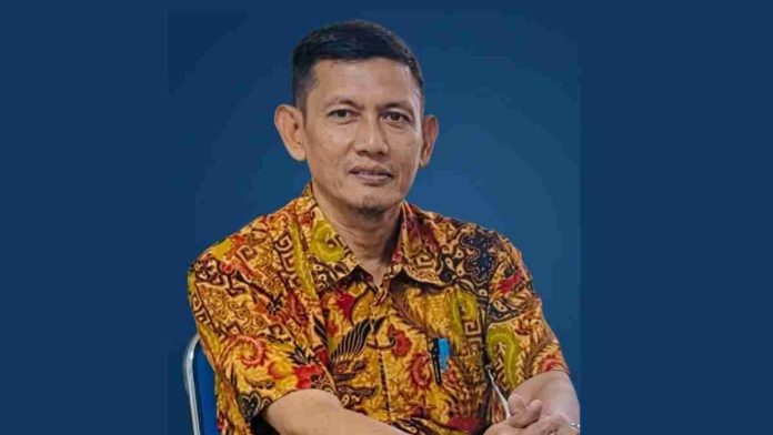 Foto potret Dedi Irawan mengenakan kemeja batik corak emas kecokelatan dengan latar belakang biru polos, posisi tangan saling bertumpu di atas meja.