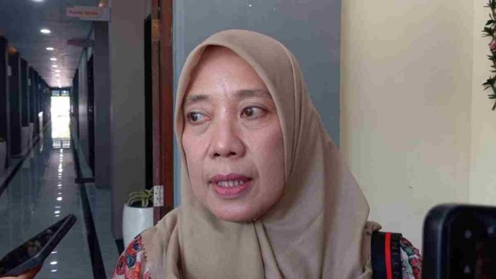 Close-up wajah Lale Widiahning mengenakan hijab cokelat sedang berbicara memberikan keterangan pers kepada wartawan di lorong sebuah gedung.