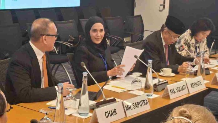 Hj. Sari Yuliati berhijab hitam duduk di meja rapat bersama delegasi internasional, memegang dokumen di depan papan nama bertuliskan Ms. Yuliati Keynote.