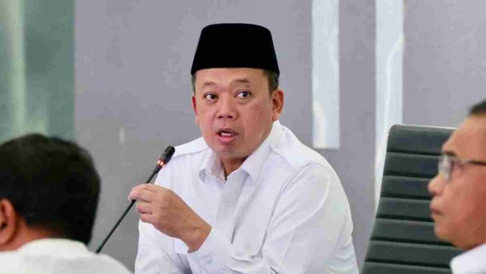 Foto close-up Menteri Nusron Wahid mengenakan kemeja putih dan peci hitam sedang berbicara serius sambil memegang mik dalam sebuah rapat pimpinan.