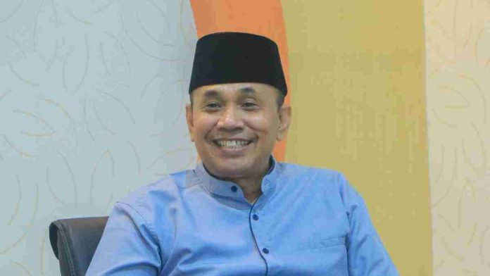 Potret Dr. H. Ahsanul Khalik mengenakan kemeja biru dan peci hitam, tampak tersenyum ramah saat memberikan keterangan di dalam ruangan.