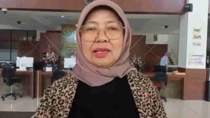 Foto Baiq Isvie Rupaeda mengenakan kemeja hitam dan blazer motif macan tutul dengan jilbab cokelat muda serta kacamata, berdiri di depan meja pelayanan kantor.
