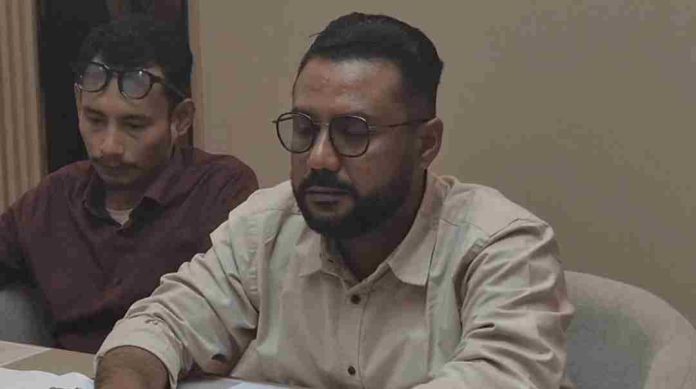 Foto seorang pria berkacamata dan berjenggot mengenakan kemeja krem, Fuad, S.H., sedang menunduk serius saat memberikan keterangan pers dalam sebuah ruangan.