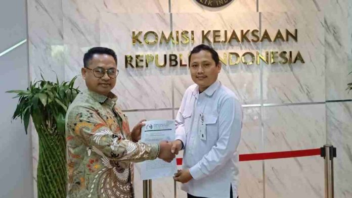 Dua pria berdiri bersalaman sambil memegang dokumen surat laporan resmi di depan dinding marmer bertuliskan Komisi Kejaksaan Republik Indonesia dengan logo emas.