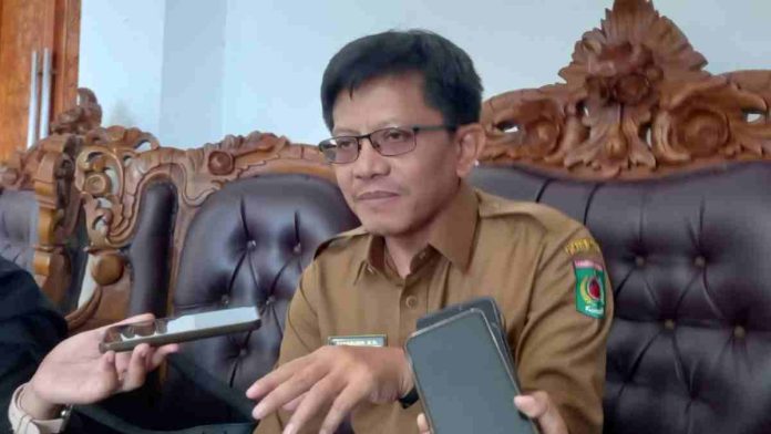 Foto Sekda Lombok Utara Sahabudin mengenakan seragam dinas cokelat dan kacamata, sedang memberikan keterangan di depan jurnalis yang memegang ponsel.