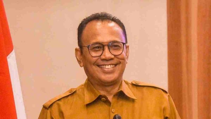 Foto potret Dr. H. Ahsanul Khalik, Kepala Dinas Kominfotik NTB, mengenakan seragam dinas cokelat dan kacamata sambil tersenyum ramah di depan latar belakang kayu.