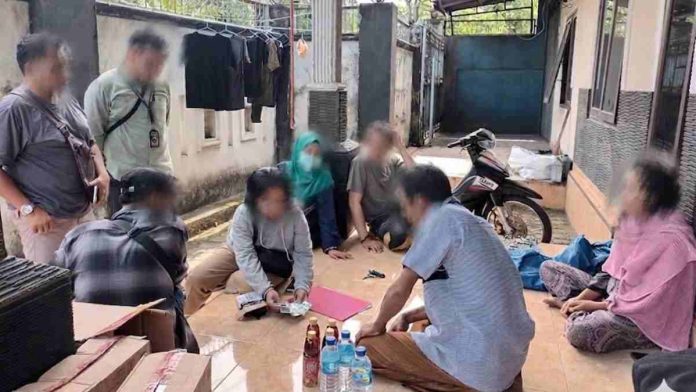 Petugas BBPOM dan kepolisian sedang memeriksa barang bukti paket obat-obatan di teras sebuah rumah bersama beberapa orang yang duduk di lantai.
