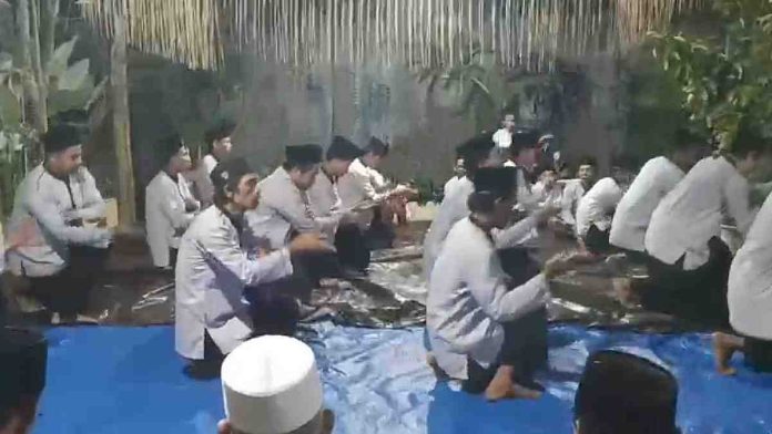 Sekelompok pria mengenakan baju koko putih dan peci hitam duduk bersimpuh di atas terpal biru dalam sebuah prosesi zikir tradisional di malam hari.