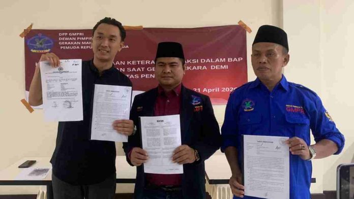 Tiga pria berdiri menunjukkan lembaran dokumen resmi di depan spanduk DPP GMPRI. Pria di kiri mengenakan kemeja hitam, tengah jas merah, dan kanan kemeja biru.