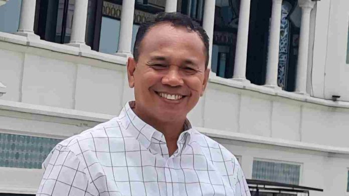 Foto potret Dr. H. Ahsanul Khalik mengenakan kemeja motif kotak-kotak putih sedang tersenyum dengan latar belakang bangunan gedung bertiang putih besar.