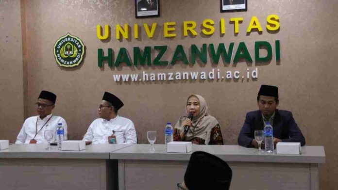 Empat orang duduk di meja panjang saat konferensi pers. Seorang wanita berhijab di tengah berbicara memegang mikrofon, didampingi tiga pria berpeci. Latar belakang dinding tertulis UNIVERSITAS HAMZANWADI.