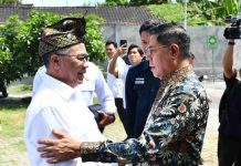Ketua Baznas RI Puji Kemandirian Lombok Timur Bangun Rumah Sakit dari Dana Zakat Masyarakat Bupati Lombok Timur mengenakan kemeja putih dan udeng tradisional bersalaman dengan pria berkacamata yang memakai kemeja batik bermotif floral di area terbuka.