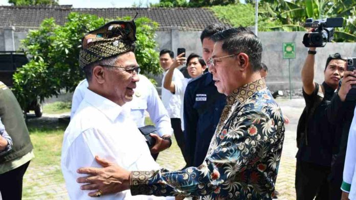 Bupati Lombok Timur mengenakan kemeja putih dan udeng tradisional bersalaman dengan pria berkacamata yang memakai kemeja batik bermotif floral di area terbuka.