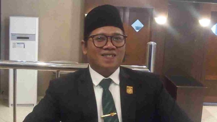 Foto potret Zia Urrahman mengenakan setelan jas hitam, kemeja putih, dasi hijau bermotif, dan peci hitam sedang tersenyum ke arah kamera dalam ruangan.