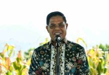 Ketua Baznas RI Puji Strategi Tani Bupati Lotim di Lahan Kering Foto potret Bupati Lombok Timur, H. Haerul Warisin, sedang tersenyum dan berbicara di depan mikrofon mengenakan kemeja batik bermotif biru. Latar belakangnya adalah ladang tanaman jagung yang menguning di bawah sinar matahari.