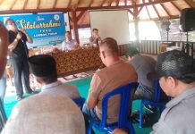 Gelar Silaturahmi Purna Tugas 88 Kades, FKKD Lotim Dorong Pilkades 2026 Foto diambil dari sudut pandang peserta di ruangan beratap kayu, menunjukkan narasumber berbicara di depan mikrofon di meja depan. Peserta duduk di kursi biru mendengarkan. Spanduk biru bertuliskan 'Silaturrahmi FKKD' terlihat jelas.