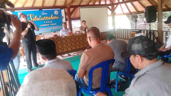Foto diambil dari sudut pandang peserta di ruangan beratap kayu, menunjukkan narasumber berbicara di depan mikrofon di meja depan. Peserta duduk di kursi biru mendengarkan. Spanduk biru bertuliskan 'Silaturrahmi FKKD' terlihat jelas.