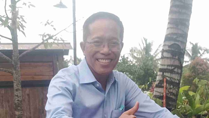 Foto potret H. Muhammad Juaini Taofik mengenakan kemeja biru muda, berkacamata, sedang tersenyum lebar dan memberikan jempol dengan latar belakang pohon kelapa.