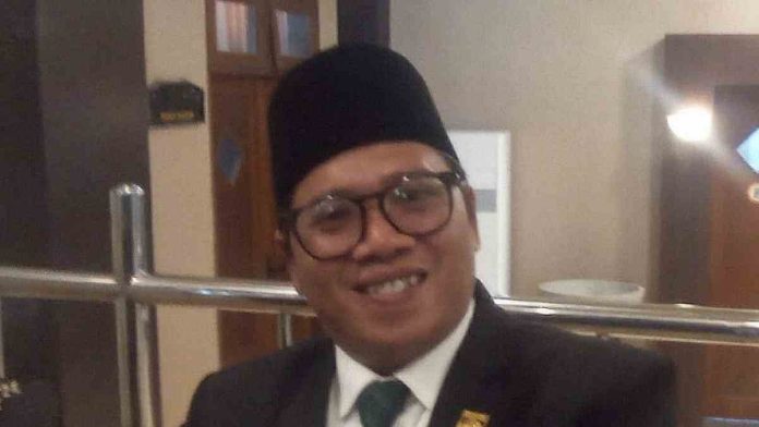 Foto potret Zia Urrahman mengenakan setelan jas hitam, kemeja putih, dasi hijau bermotif, dan peci hitam sedang tersenyum ke arah kamera dalam sebuah ruangan formal.