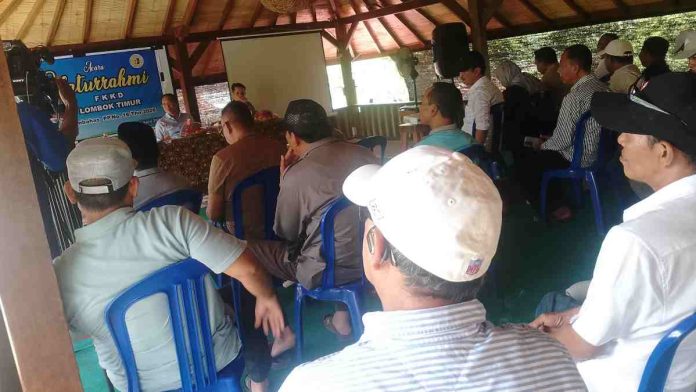 Suasana pertemuan di aula terbuka beratap kayu (gazebo besar). Peserta yang merupakan pria mengenakan topi dan kemeja duduk membelakangi kamera menghadap narasumber.