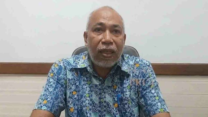 Foto potret Muhammad Taswiruddin mengenakan kemeja batik biru bermotif kuning dan putih, memiliki jenggot putih, sedang berbicara menghadap kamera dengan latar dinding putih.