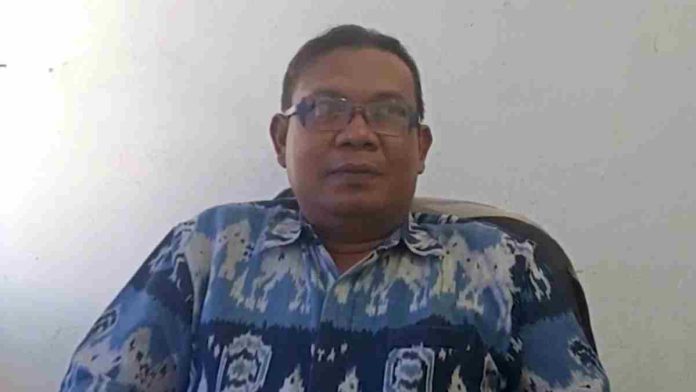 Foto potret Mirza Sopian mengenakan kemeja kain tenun ikat berwarna biru motif etnik, berkacamata, sedang memberikan keterangan dengan latar belakang dinding putih.