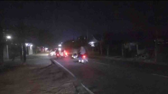 Pemandangan jalan raya aspal pada malam hari yang gelap dengan beberapa pengendara sepeda motor sedang melintas, lampu kendaraan menjadi sumber cahaya utama.
