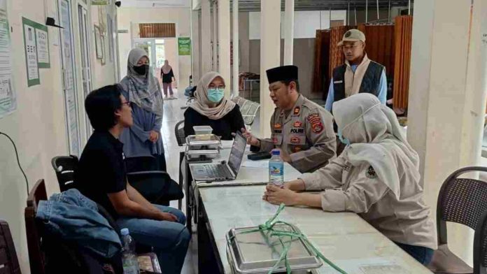 Seorang anggota polisi berseragam dinas duduk di meja pendaftaran puskesmas sedang berdialog dengan petugas kesehatan dan warga, didampingi unit intelkam yang mendata korban.