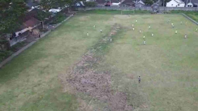 Foto udara lapangan sepak bola dengan rumput hijau yang tidak merata, menunjukkan beberapa titik tanah gersang dan pemain bola sedang beraktivitas di lapangan.