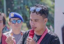 IMM Desak Bupati Lotim Kembalikan Camat dan Sekcam Siaga di Pringgabaya Seorang pemuda berkaos hitam dengan kacamata hitam di atas kepala sedang berbicara serius memegang mikrofon merah, didampingi aparat kepolisian di latar belakang.