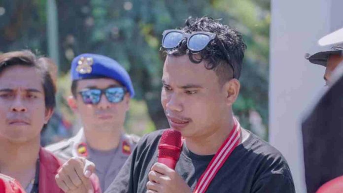 Seorang pemuda berkaos hitam dengan kacamata hitam di atas kepala sedang berbicara serius memegang mikrofon merah, didampingi aparat kepolisian di latar belakang.