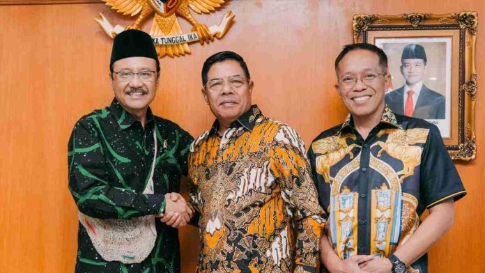 Tiga orang pria berdiri sejajar di depan lambang Garuda Pancasila. Bupati Haerul Warisin berbaju batik hijau bersalaman dengan Mensos Gus Ipul, didampingi Sekda Lotim.