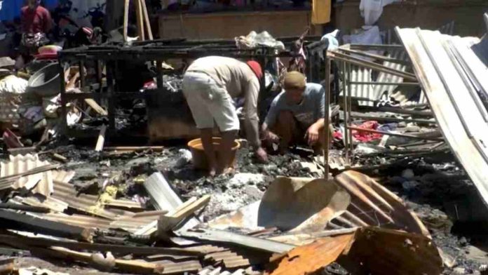 Dua orang pria berbaju santai sedang membungkuk mengais sisa-sisa barang di tengah puing bangunan pasar yang hancur dan menghitam akibat api.