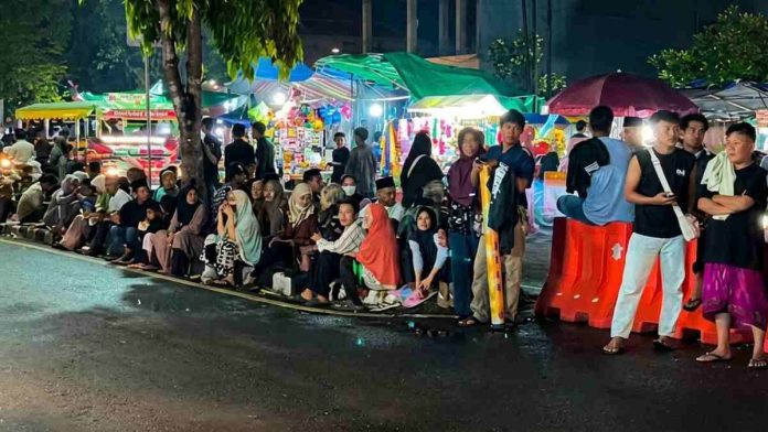 Pemandangan malam hari memperlihatkan kerumunan warga duduk berjejer di trotoar pinggir jalan raya depan area asrama haji yang ramai dengan pedagang kaki lima.